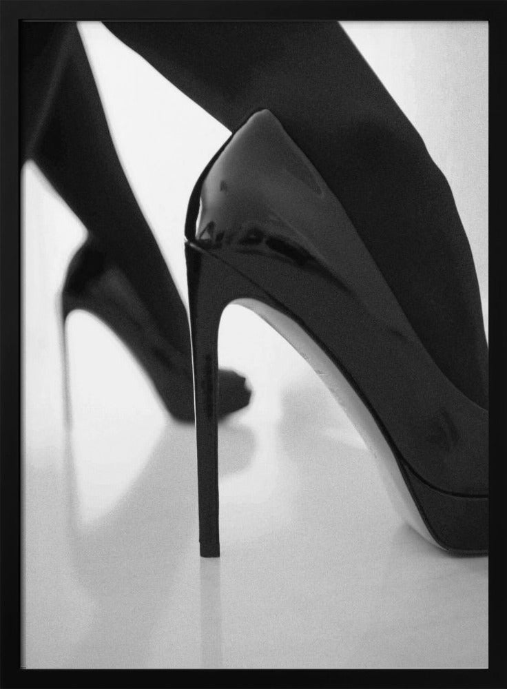 A close-up, black and white photograph of a person wearing shiny black patent leather stilettos and dark stockings. One shoe is in sharp focus in the foreground, highlighting its high arch and thin heel, while the other leg and shoe are softly blurred in the background on a reflective white surface. Decor