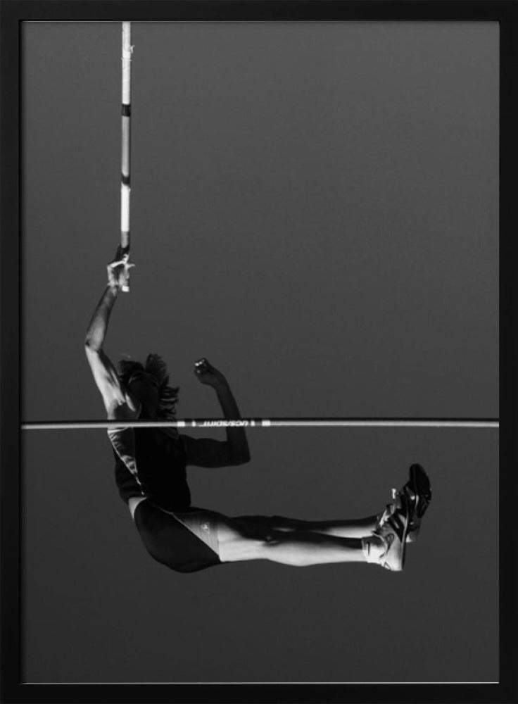 A dramatic black and white photograph of a male pole vaulter in mid-air, successfully clearing the horizontal bar. The athlete's body is parallel to the ground, with one arm still holding the vertical pole, against a solid gray sky. The image is enclosed in a simple black frame. Artwork