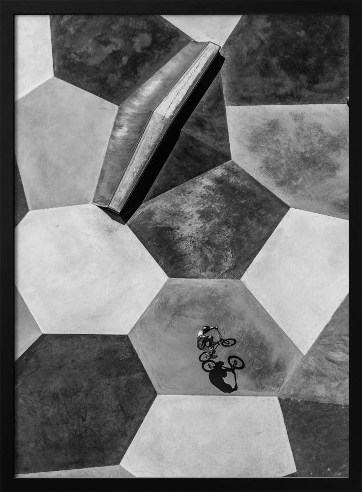A high-angle, black and white photograph of a BMX rider in a skatepark with a geometric pattern. The ground consists of large, interlocking hexagons in various shades of grey, and the rider is captured mid-action on one of them, with a sharp shadow cast beside them. Artwork