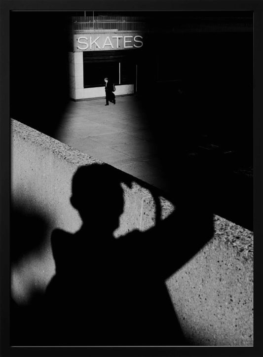 A high-contrast, black and white street photograph where the large shadow of the photographer is cast on a concrete barrier in the foreground. In the background, a lone person walks through a spotlit area of sidewalk in front of a building with an illuminated sign that says 'SKATES'. Poster