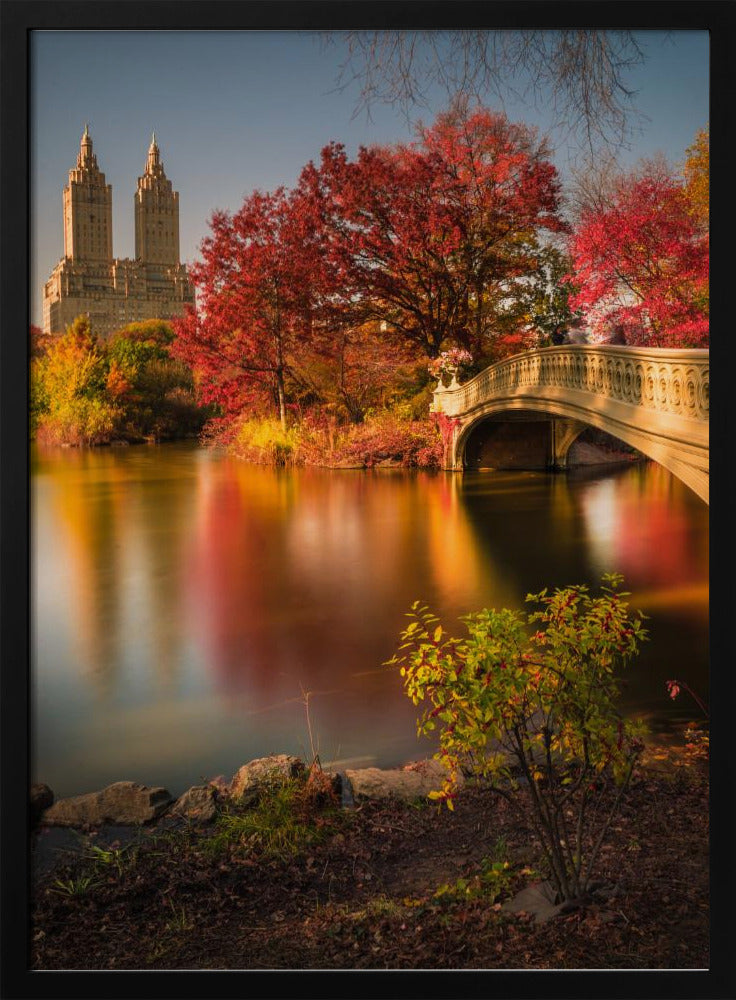 A picturesque autumn scene in Central Park featuring the Bow Bridge over a calm lake reflecting the vibrant red and yellow fall foliage, with the twin towers of The San Remo building visible in the background against a clear blue sky. Decor