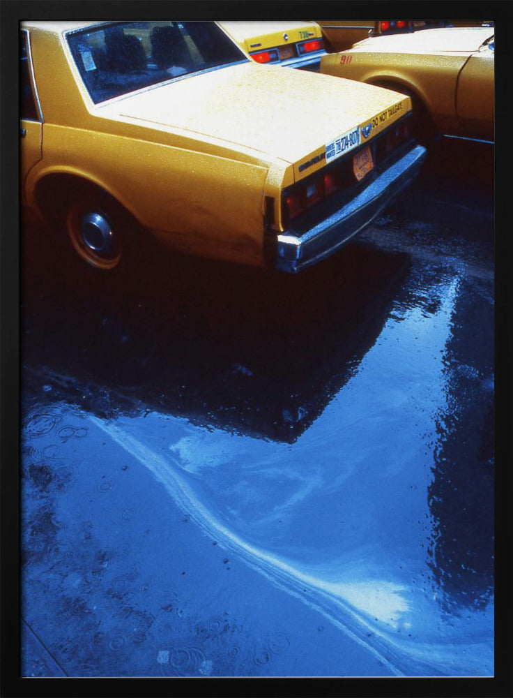 A close-up, angled view of the rear of a yellow taxi cab on a wet street. The car's dark reflection is cast upon a large, vibrant blue puddle in the foreground, with visible ripples from the rain. Wall Art