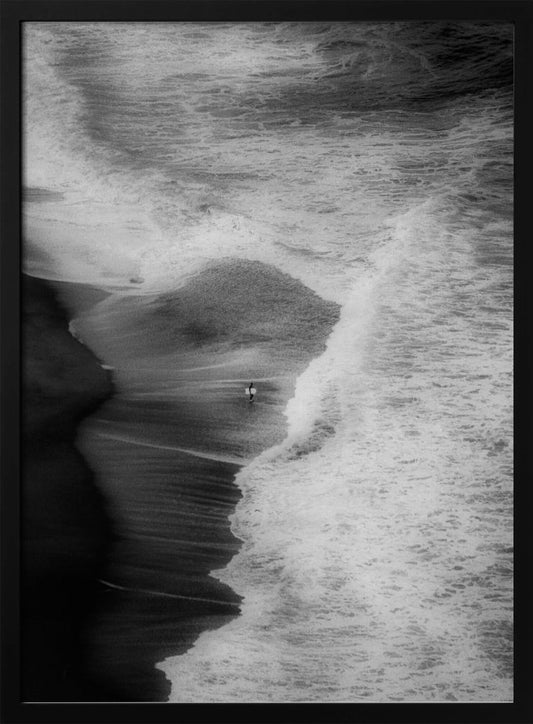 A high-angle, black and white photograph of a lone surfer carrying a surfboard at the water's edge on a dark sand beach. The vast, textured ocean with large, foamy waves crashing ashore dwarfs the solitary figure, creating a sense of scale and solitude. Decor
