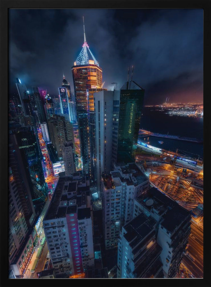 A dramatic high-angle nighttime photograph of a sprawling modern cityscape, with towering skyscrapers illuminated in vibrant blue, orange, and purple neon lights under a dark, cloudy sky. Below, streets are aglow with traffic, and in the distance, a brightly lit harbor meets the dark water. Decor