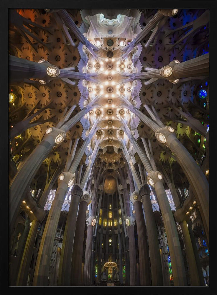 A low-angle, symmetrical photograph looking up at the incredibly ornate and complex vaulted ceiling of the Sagrada Familia Basilica. The massive stone columns branch out like trees, supporting the intricately detailed ceiling illuminated by warm, glowing lights and splashes of color from unseen stained glass windows. Poster