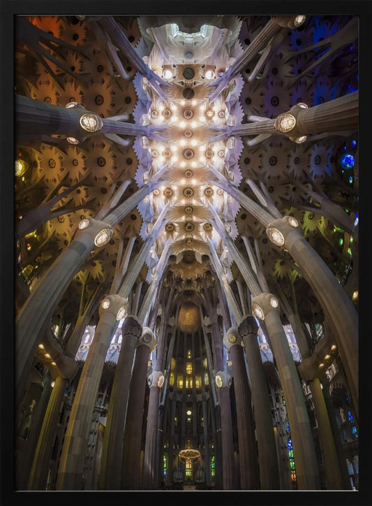 A low-angle, symmetrical photograph looking up at the incredibly ornate and complex vaulted ceiling of the Sagrada Familia Basilica. The massive stone columns branch out like trees, supporting the intricately detailed ceiling illuminated by warm, glowing lights and splashes of color from unseen stained glass windows. Poster