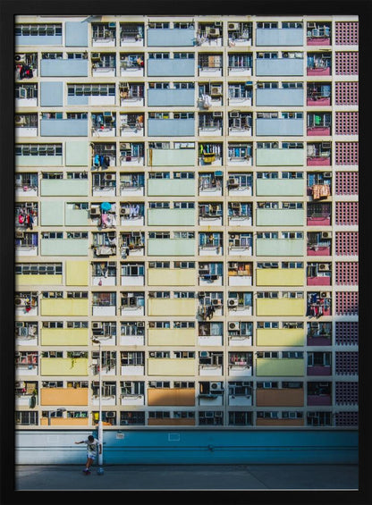 A symmetrical, full-frame shot of a massive apartment building with a repeating grid pattern of windows and balconies. The building is painted in horizontal bands of pastel colors, starting with blue at the top, then green, yellow, and orange. A single boy plays with a soccer ball on the ground level in front of a blue wall. Wall Art