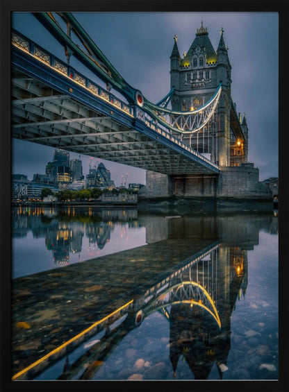 A dramatic, low-angle photograph of London's Tower Bridge at dusk. The iconic bridge is illuminated with warm yellow lights against a dark blue, cloudy sky. The calm River Thames in the foreground creates a stunning, clear reflection of the bridge and the distant city skyline. Print
