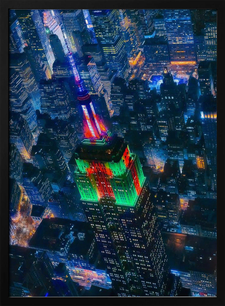 A dramatic high-angle aerial view of the Empire State Building at night, illuminated in festive red and green lights. The surrounding New York City skyline is a dense grid of skyscrapers glowing with blue and white lights, creating a vibrant, sprawling cityscape. Poster