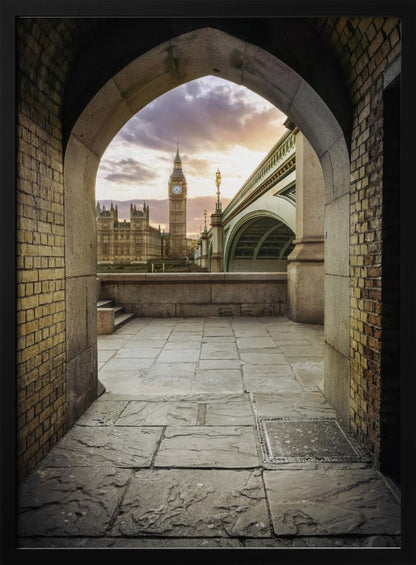 A scenic view of London's Big Ben clock tower and the Houses of Parliament as seen through a weathered stone archway. The foreground features a stone-paved walkway, and the background shows the iconic landmarks against a cloudy sunset sky with hues of purple and orange. Poster
