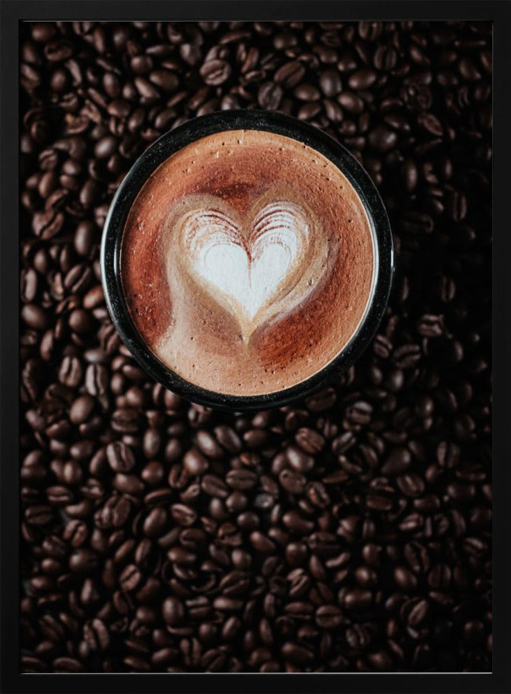 A top-down view of a cappuccino in a black mug with a white heart-shaped foam art design, placed on a bed of dark roasted coffee beans. Artwork