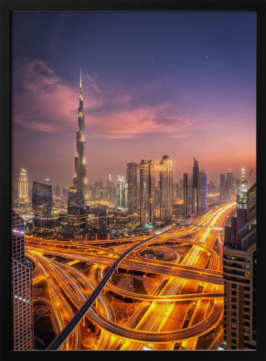 A stunning aerial photograph of the Dubai skyline at dusk, featuring the illuminated Burj Khalifa against a purple and pink sunset sky. In the foreground, complex highway interchanges are aglow with the golden-orange light trails of moving traffic, creating a sense of vibrant energy. Poster