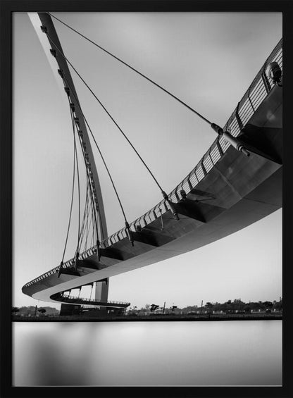 A dynamic low-angle, black and white photograph of a modern cable-stayed pedestrian bridge. The bridge curves elegantly over calm, reflective water, with its deck suspended by numerous cables attached to a tall, angled pylon, creating a striking graphic composition against a bright, clear sky. Decor