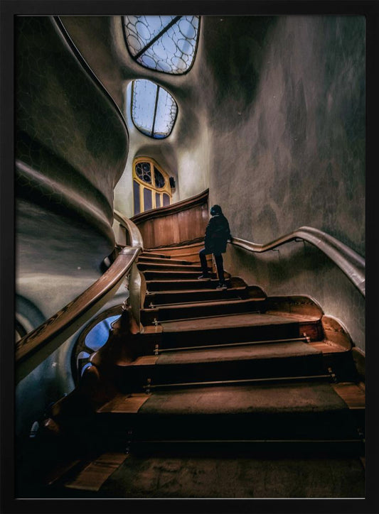 A low-angle view up a dark, winding wooden staircase inside a building with surreal, organic architecture. A person in a dark coat is walking up the stairs towards uniquely shaped windows that show a blue sky, contrasting with the dimly lit, textured gray walls. Print