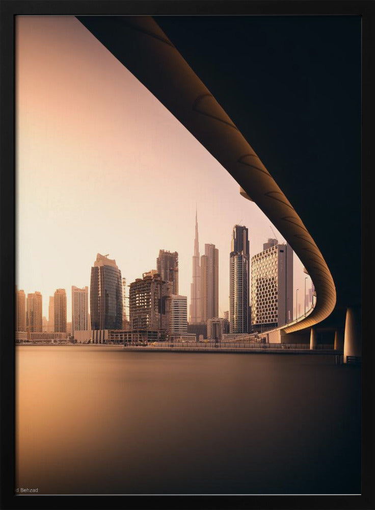 A view of the Dubai skyline, including the Burj Khalifa, framed by the sweeping curve of a bridge in the foreground. The city is bathed in the warm, soft light of a sunrise or sunset, with its reflection visible on the calm water below. Decor