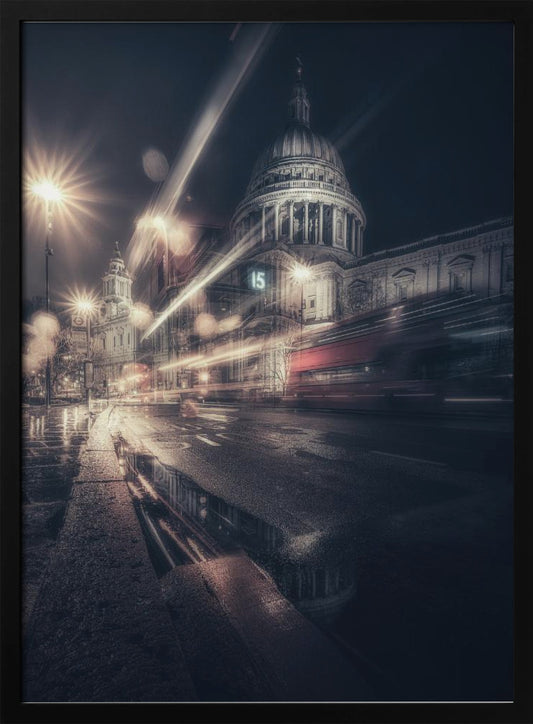 A dynamic long-exposure photograph of a wet London street at night, with the iconic dome of St. Paul's Cathedral illuminated in the background. Streaks of light from passing traffic, including a red bus, blur across the scene, while the glistening pavement reflects the cathedral and bright streetlights. Decor