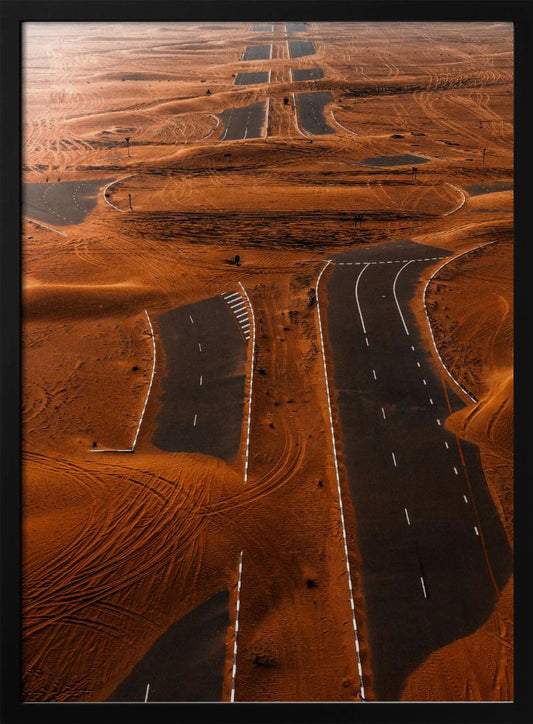 An aerial view of a multi-lane, black asphalt road being consumed by vast, rolling sand dunes of a deep orange color. The sand covers sections of the road, creating a surreal pattern of man-made lines and natural, windswept textures. Print