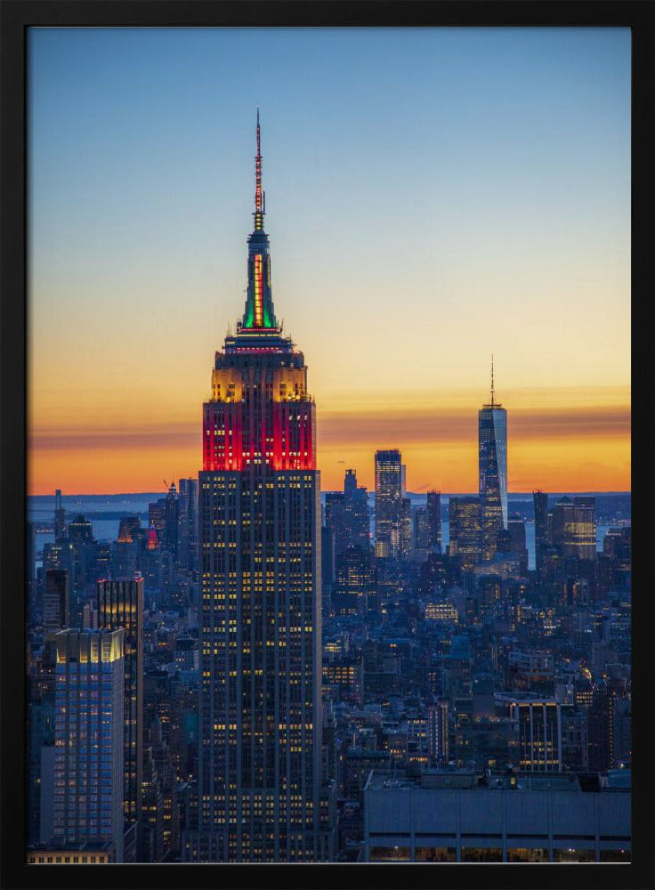 A vertical photograph of the New York City skyline at sunset, with the Empire State Building in the foreground. The building is illuminated with festive red and green lights on its upper floors, set against a sky that transitions from deep blue to a warm orange glow at the horizon. The surrounding cityscape, including the distant One World Trade Center, is bathed in the cool light of dusk, with scattered warm lights from building windows. Decor