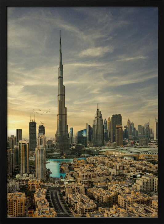 A framed photograph of the Dubai skyline at sunset, with the iconic Burj Khalifa skyscraper dominating the center. Surrounding it are numerous other modern high-rise buildings, a body of turquoise water, and a dense urban landscape in the foreground, all under a cloudy, golden-hued sky. Poster