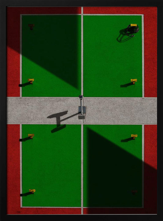 A high-angle, symmetrical photograph of a green and red sports court, viewed from directly above. Strong, sharp shadows cut diagonally across the composition, creating a geometric and abstract pattern of light and dark. A net post in the center casts a T-shaped shadow on the concrete dividing strip. Print