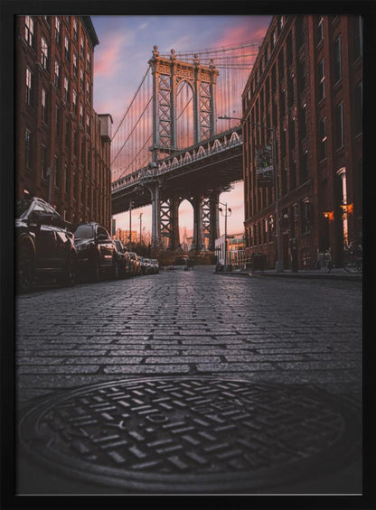 A low-angle photograph taken from a cobblestone street in Dumbo, Brooklyn, looking up towards the Manhattan Bridge at sunset. Red brick buildings frame the view on both sides of the street, with parked cars lining the left. A manhole cover is prominent in the foreground, and the sky is filled with warm pink and orange tones. Print