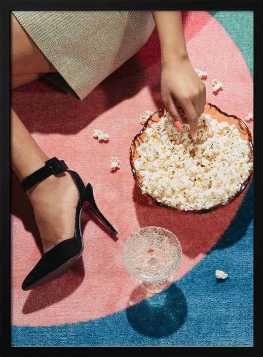 A high-angle photograph of a woman sitting on a vibrant pink and blue rug. She is wearing a chic black stiletto high heel and reaching into a glass bowl filled with popcorn. A crystal glass and scattered popcorn kernels are also on the rug. Artwork