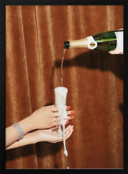 Champagne is poured from a bottle into a glass, overflowing with white foam that drips down onto two manicured hands with red nail polish holding it against a brown velvet curtain background. Poster