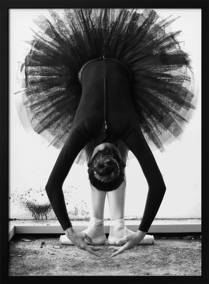 A striking black and white photograph of a ballerina in a deep forward bend. Her body is clad in a black leotard and white tights, with a voluminous black tutu fanning out vertically behind her. Her hair is tied in a bun, and her hands gracefully reach towards her feet, which are in white pointe shoes. Artwork