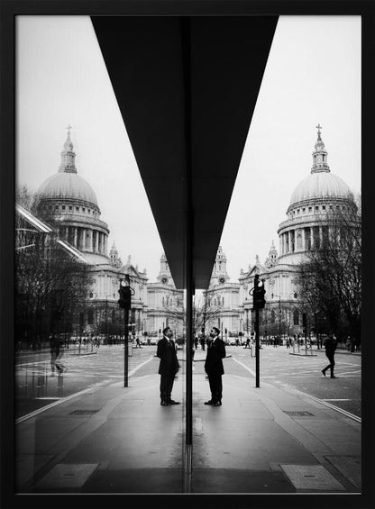 A surreal black and white photograph capturing a symmetrical reflection of a city scene, with St. Paul's Cathedral prominently featured on both sides. A dark, V-shaped reflection from a building divides the image vertically. In the foreground, the reflection of two men in dark suits standing on the sidewalk adds a human element to the grand architectural view. Decor