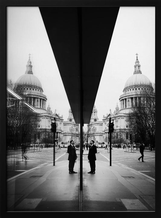 A surreal black and white photograph capturing a symmetrical reflection of a city scene, with St. Paul's Cathedral prominently featured on both sides. A dark, V-shaped reflection from a building divides the image vertically. In the foreground, the reflection of two men in dark suits standing on the sidewalk adds a human element to the grand architectural view. Decor