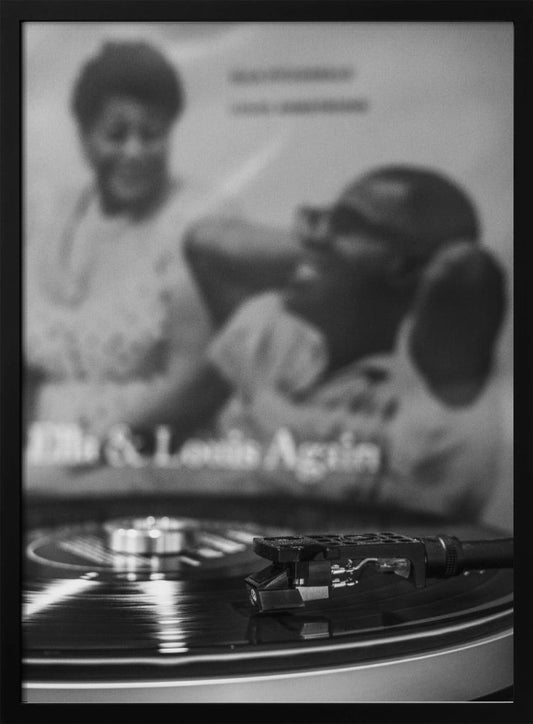 A black and white photograph focusing on a turntable's stylus resting on a spinning vinyl record. In the blurred background, the album cover for 'Ella & Louis Again' featuring Ella Fitzgerald and Louis Armstrong is visible. Poster