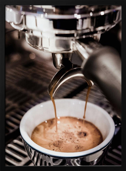 A close-up photograph of a shiny chrome espresso machine brewing a fresh shot of espresso. Two streams of rich, dark coffee pour from the portafilter into a white ceramic cup with a blue patterned rim, creating a light brown crema on top. Print