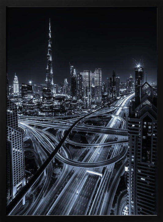 A cool-toned black and white photograph of the Dubai skyline at night. The iconic Burj Khalifa stands tall in the background, while the foreground is dominated by a massive, complex highway interchange with light trails from moving cars, creating a sense of motion and modernity. Poster
