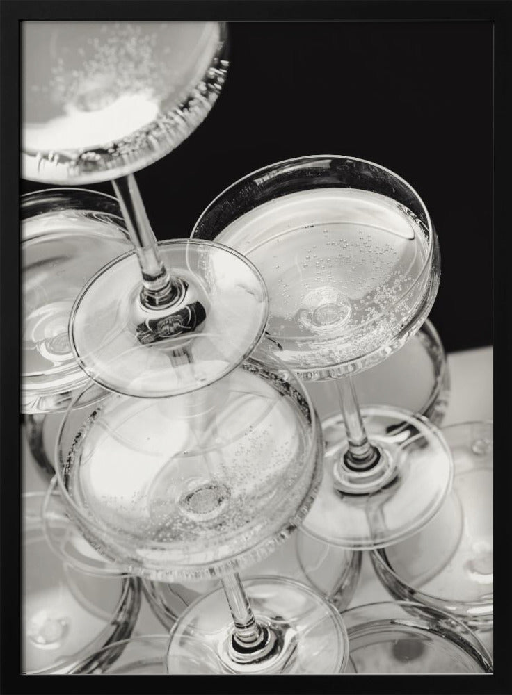 A close-up, high-angle photograph in black and white of a champagne tower. Several coupe-style glasses are stacked, filled with bubbly champagne that catches the light. The background is dark, creating a strong contrast and highlighting the elegant glassware. Decor