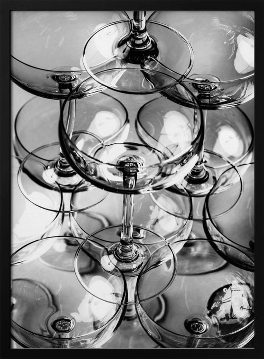 A black and white, overhead photograph of a tightly packed stack of empty champagne coupe glasses. The overlapping rims, stems, and bowls create a complex abstract pattern of circles and reflections, all enclosed within a simple black frame. Poster