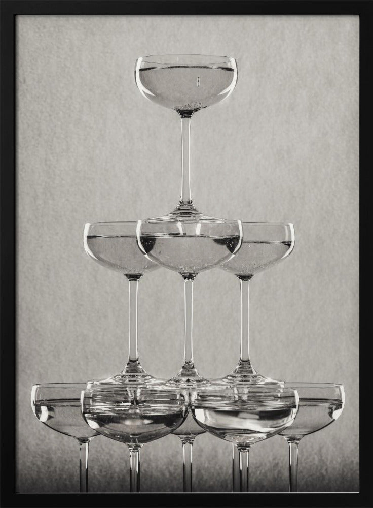 A classic black and white photograph of a champagne tower, with coupe glasses stacked in a pyramid. The glasses are filled with bubbly liquid, and the composition is set against a textured light gray background and enclosed in a black frame. Poster