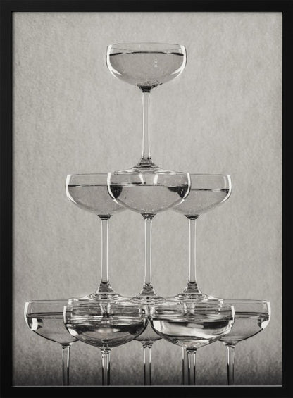 A classic black and white photograph of a champagne tower, with coupe glasses stacked in a pyramid. The glasses are filled with bubbly liquid, and the composition is set against a textured light gray background and enclosed in a black frame. Poster