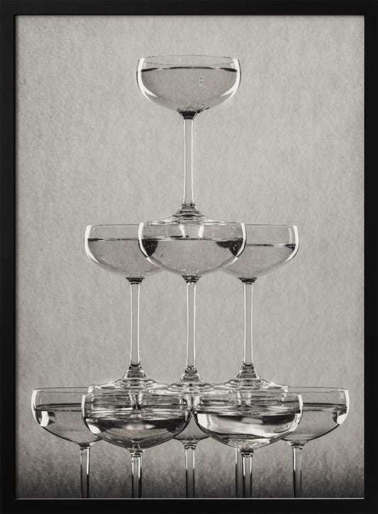 A classic black and white photograph of a champagne tower, with coupe glasses stacked in a pyramid. The glasses are filled with bubbly liquid, and the composition is set against a textured light gray background and enclosed in a black frame. Poster