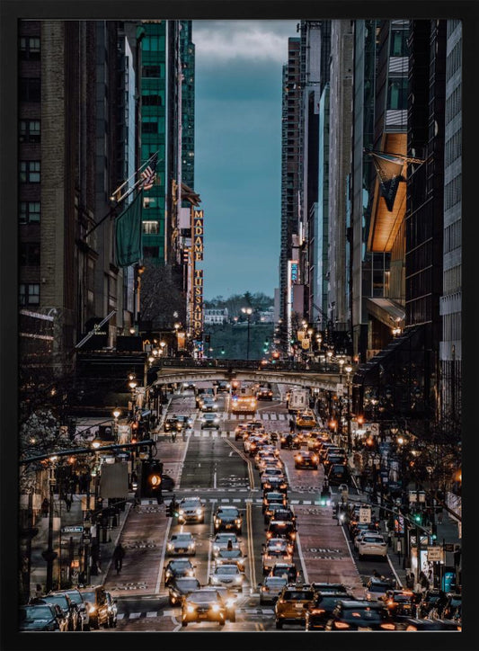 A high-angle, vertical photograph looking down a bustling New York City street at dusk, framed by towering skyscrapers. The street below is packed with cars, their headlights creating a warm golden glow, contrasting with the cool, dark blue tones of the twilight sky and buildings. Poster