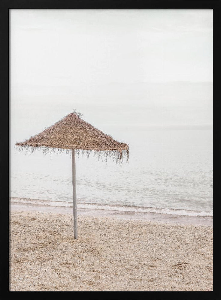 A minimalist photograph of a solitary thatched umbrella on a sandy beach, with calm, grey water and a hazy, overcast sky in the background, creating a serene and tranquil coastal scene. Poster