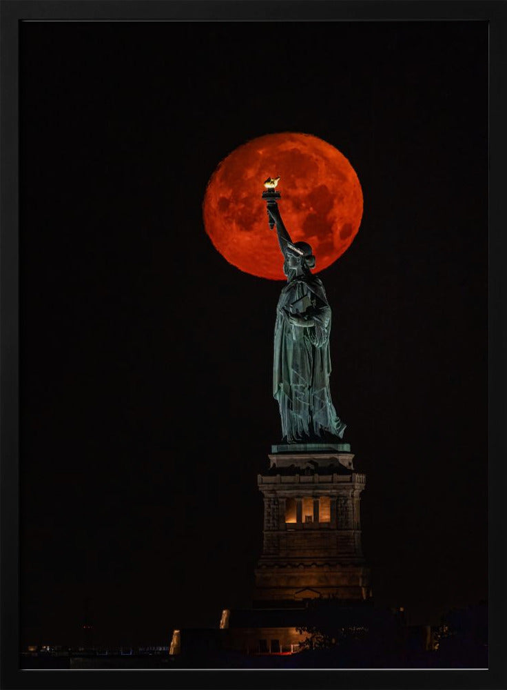 A striking night photograph of the Statue of Liberty perfectly silhouetted against a giant, vibrant red supermoon, which appears to be held aloft by the statue's torch against a deep black sky. Artwork