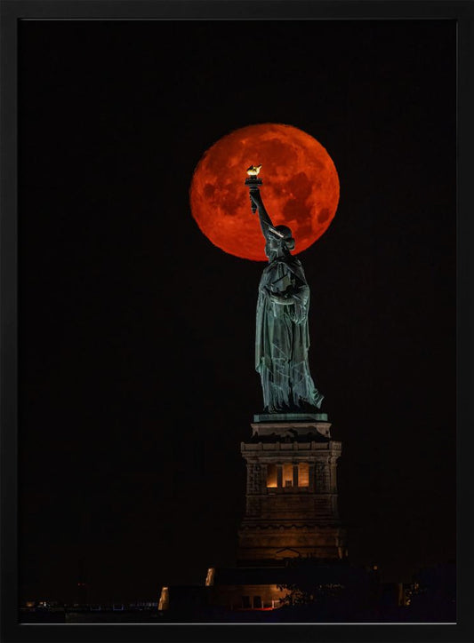 A striking night photograph of the Statue of Liberty perfectly silhouetted against a giant, vibrant red supermoon, which appears to be held aloft by the statue's torch against a deep black sky. Artwork