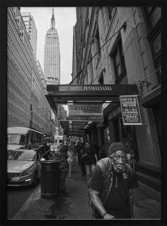 A powerful black and white vertical photograph capturing a New York City street scene. An elderly Black man with a weary expression is in the foreground, walking along a crowded sidewalk. In the background, the Empire State Building towers over the cityscape, framed between buildings, with a sign for the Hotel Pennsylvania visible. Artwork