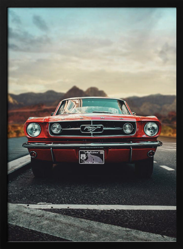 A low-angle, head-on shot of a vintage red Ford Mustang parked on an asphalt road. The iconic silver horse emblem is visible on the grille. In the background, a desert mountain range is blurred under a cloudy, muted sky. Poster