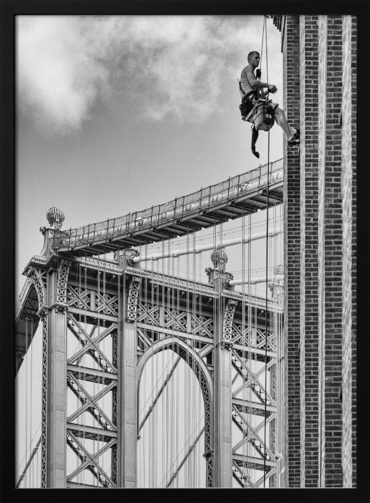 A black and white photograph capturing a window washer rappelling down the side of a tall brick building. In the background, the intricate steel structure of the Manhattan Bridge stands against a partly cloudy sky, creating a powerful urban scene. Decor
