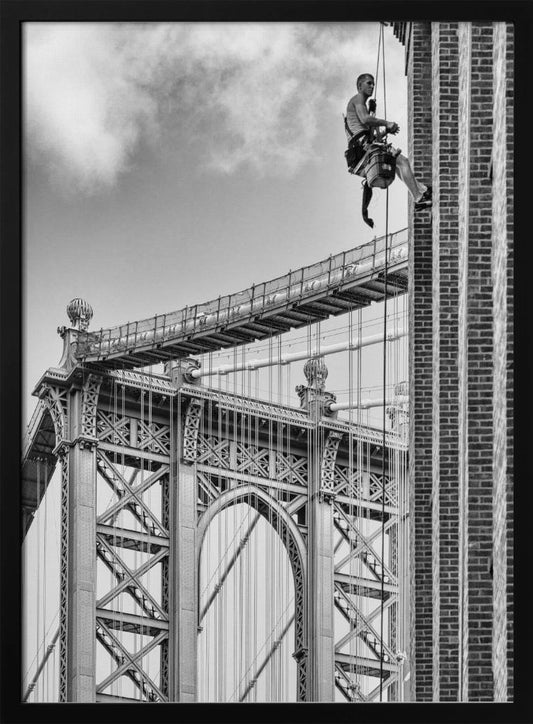 A black and white photograph capturing a window washer rappelling down the side of a tall brick building. In the background, the intricate steel structure of the Manhattan Bridge stands against a partly cloudy sky, creating a powerful urban scene. Decor