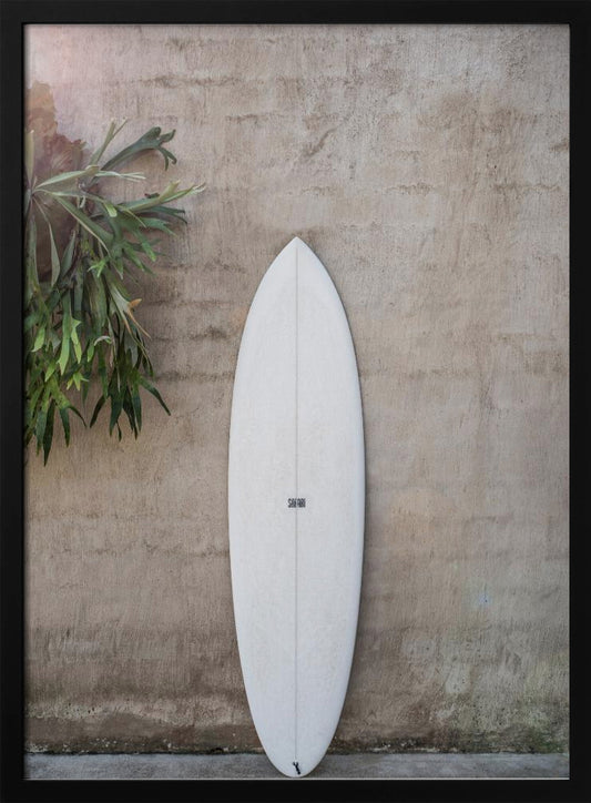 A minimalist photograph of a white surfboard leaning against a textured beige concrete wall. To the left, a large green staghorn fern adds a touch of nature. The entire scene is enclosed in a simple black frame. Artwork