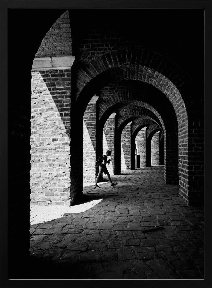 A dramatic black and white photograph of a person walking through a long brick arcade. Sunlight creates strong, contrasting shadows, highlighting the repeating arches that recede into the darkness, creating a powerful sense of depth and perspective. Decor