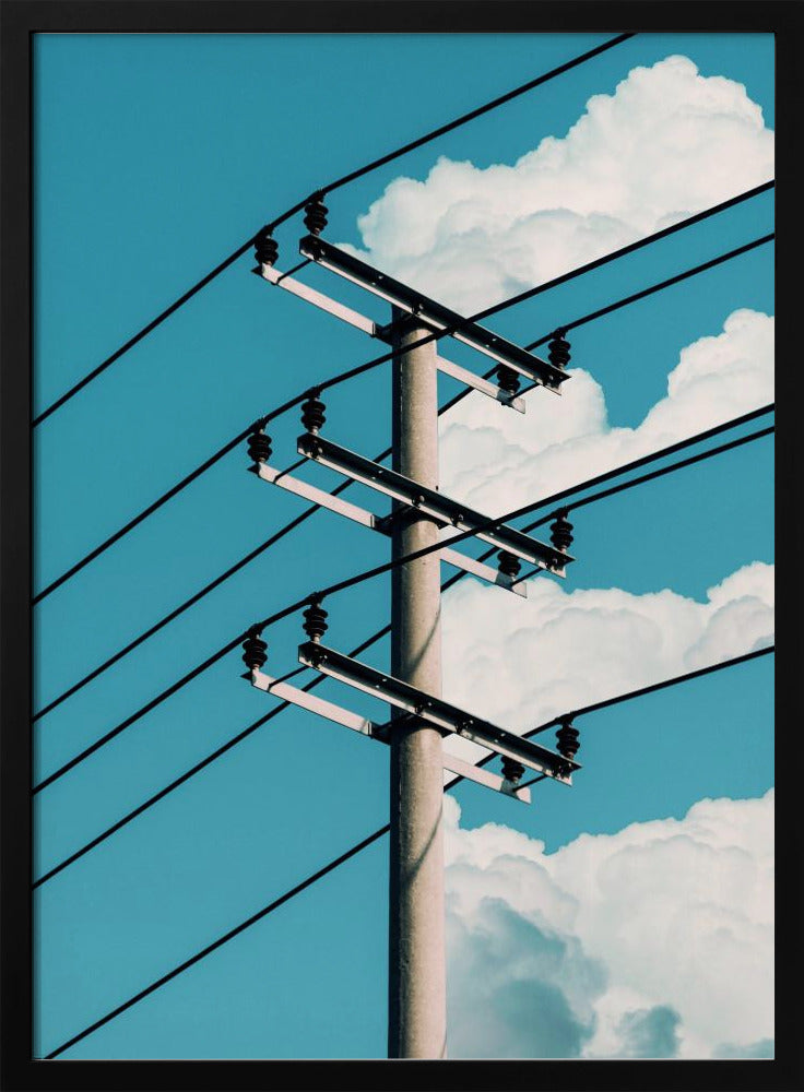 A low-angle photograph of a concrete utility pole with black power lines stretching diagonally across the frame against a vibrant blue sky with fluffy white clouds. Decor