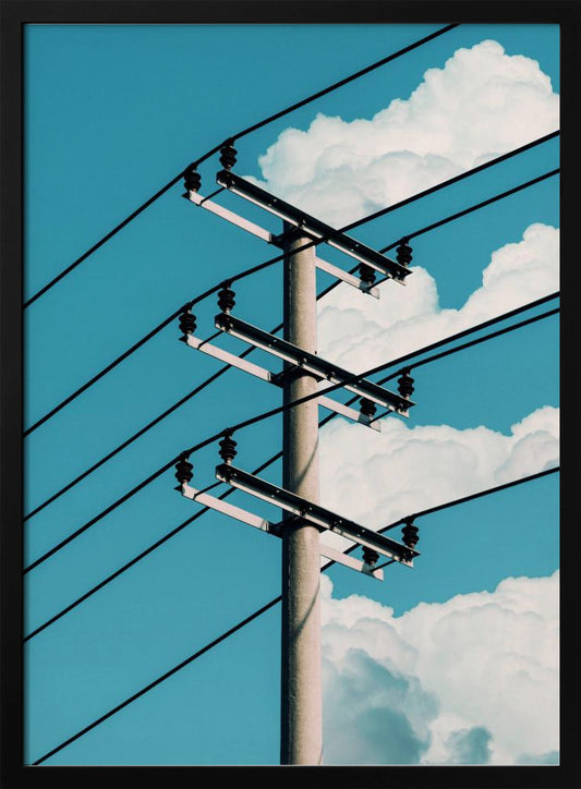 A low-angle photograph of a concrete utility pole with black power lines stretching diagonally across the frame against a vibrant blue sky with fluffy white clouds. Decor